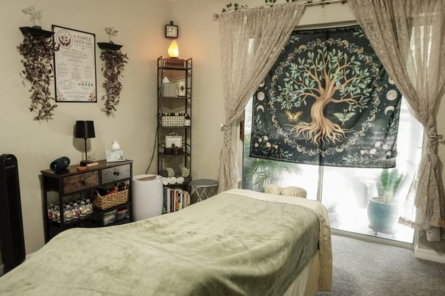 The treatment room: the massage table made up with a sage-green blanket and a bolster, a tree-of-life tapestry in the window, and a salt lamp, shelves of towels and books, and a framed copy of the welcome sheet on the wall