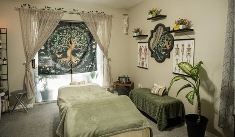 The treatment room: a massage table under a sage-green blanket, a tree-of-life tapestry hung in the window behind it, and lace curtains, anatomy charts and houseplants around the walls