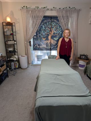 Cassandra Myers standing at the head of the massage table in the treatment room, with a tree-of-life tapestry in the window behind her